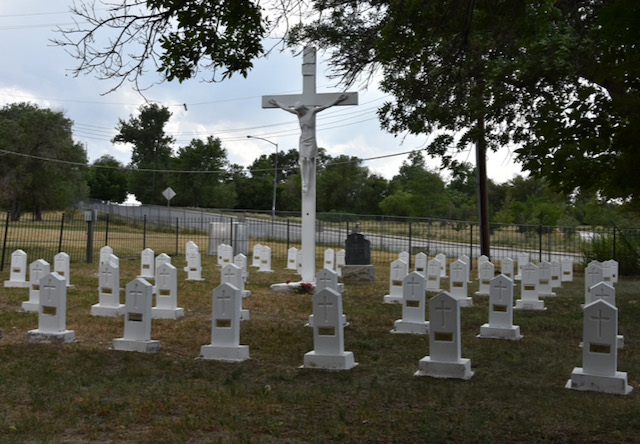 A large white crucifix stands over a cluster of white gravestones surrounded by an iron fence.