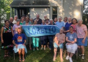 A group of people gather for a photo in front of a house around a banner that reads "Loretto."