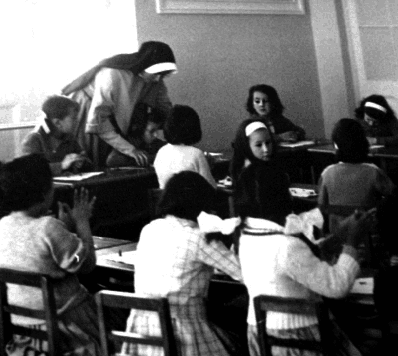 A nun in a habit helps one student while others in the classroom look on.