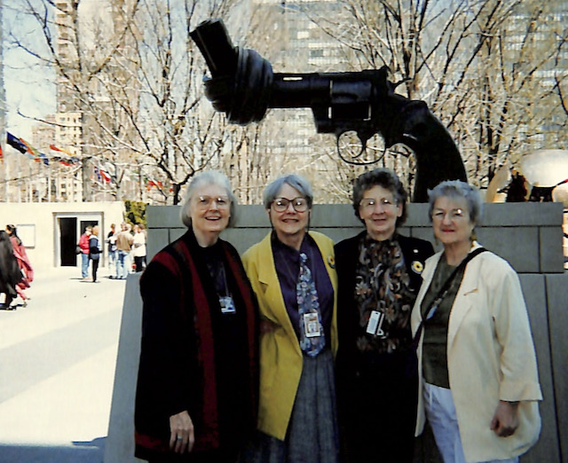 Four women pose at the base of a large statue of a gun, the barrel of which has been tied in a knot.