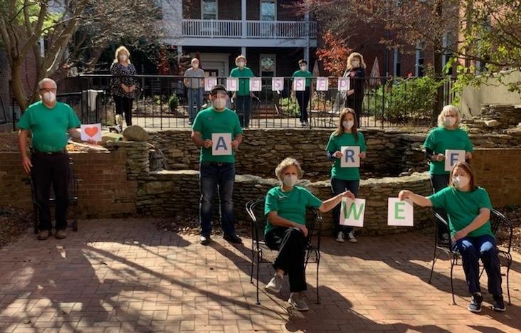 People in green shirts and N95 masks are spaced at a distance in a multi-level courtyard, holding letters on paper that spell out "WE ARE BLESSED."