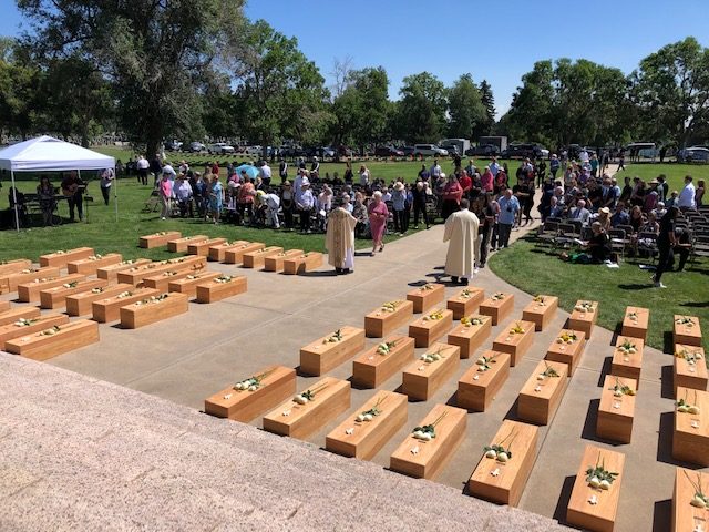 Four neat rows of wooden coffins, white roses on each, are in the foreground. An audience is visible in the background.  