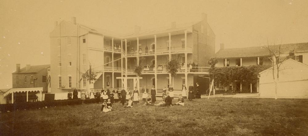 Archival photo of large, L-shaped three-story building with balconies running the length of all three floors.