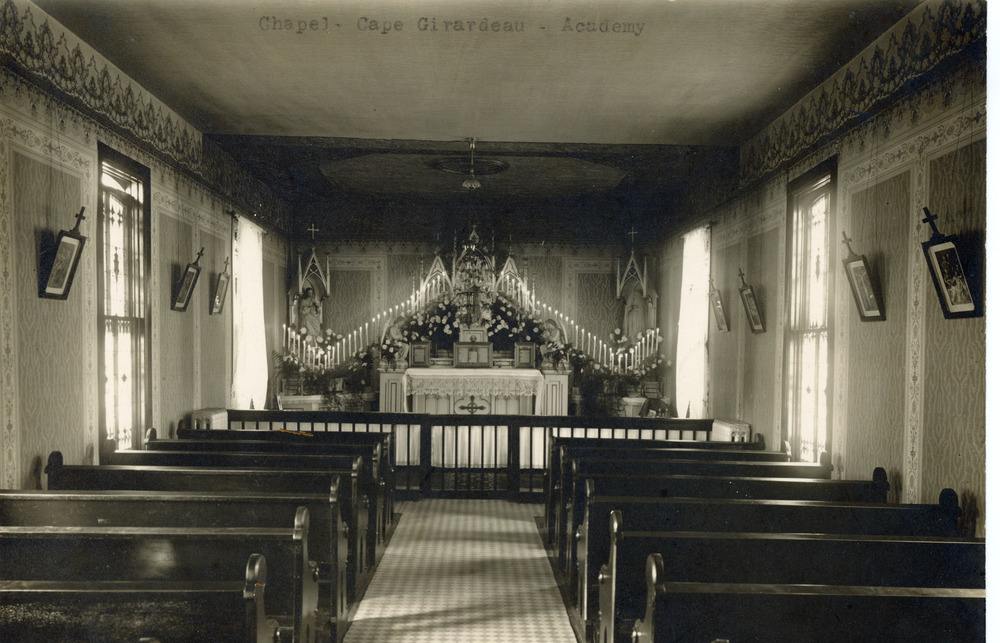 Archival photo of the inside of a chapel taken from the center aisle in the back of the chapel. The pews, communion rail and altar can be seen at the front of the church..