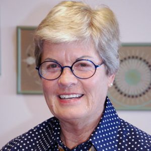A woman with short grey hair and round black-framed glasses wearing a navy blue and white polka dot collared button up and colorful patterned tie smiling for a headshot picture indoors.