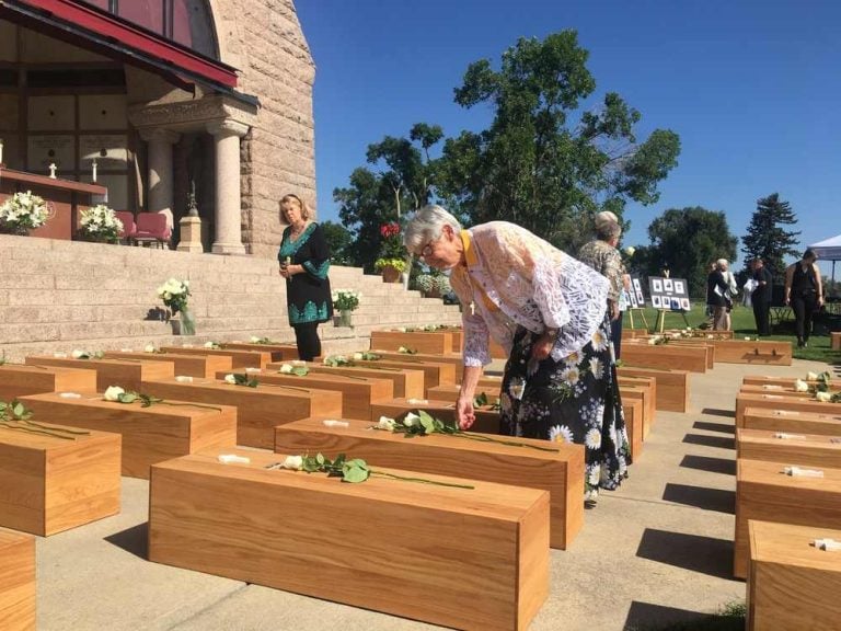 Amidst rows of wooden caskets, a woman bends down to lay a flower on top of one.
