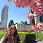 young girl smiles big and poses in front of a city background and pink blossom tree.