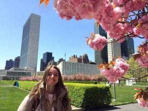 young girl smiles big and poses in front of a city background and pink blossom tree.