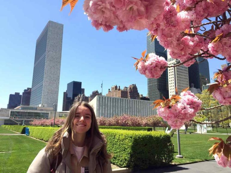 young girl smiles big and poses in front of a city background and pink blossom tree.