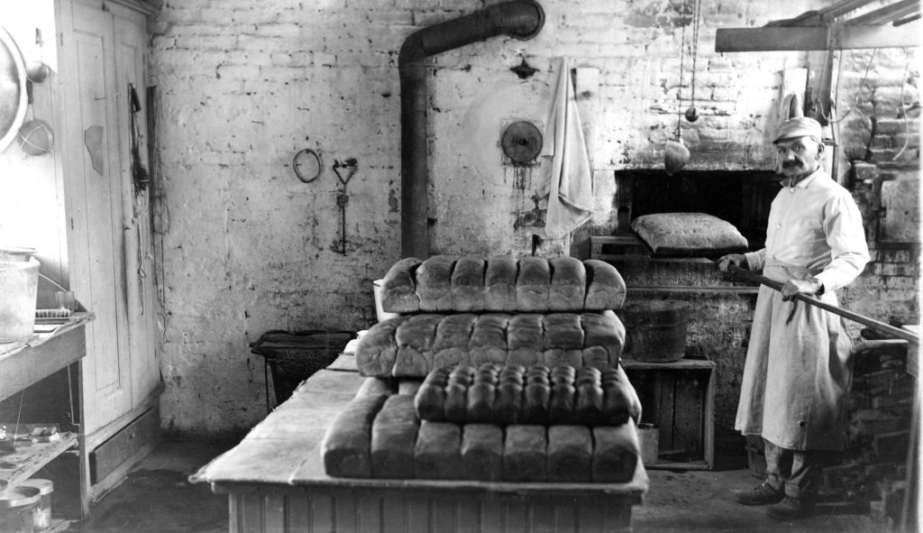 Archival photo of a mustached man in a white hat and apron standing in an old brick kitchen next to a table with stacks of bread, holding a bread paddle with multiple loaves on it.