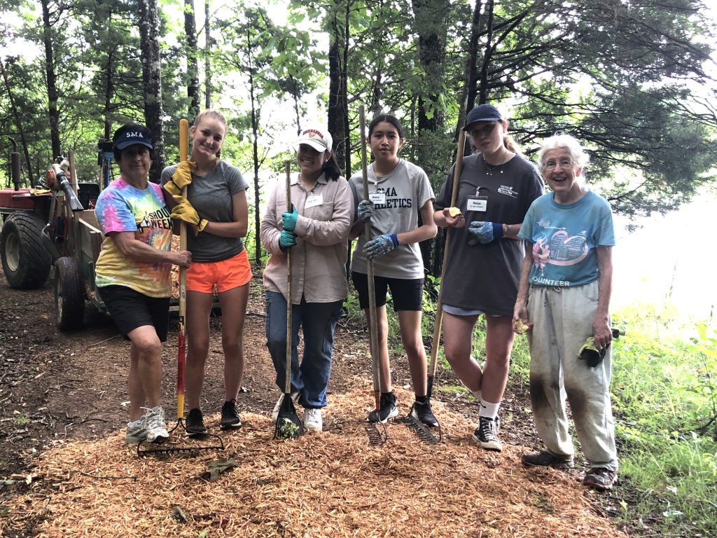 Six people stand over a pile of mulch on a trail in the woods, holding rakes and wearing gloves 