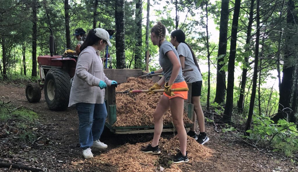 Young women raking mulch out of the back of a tractor-pulled trailer, so as to spread it on the trail in the woods.