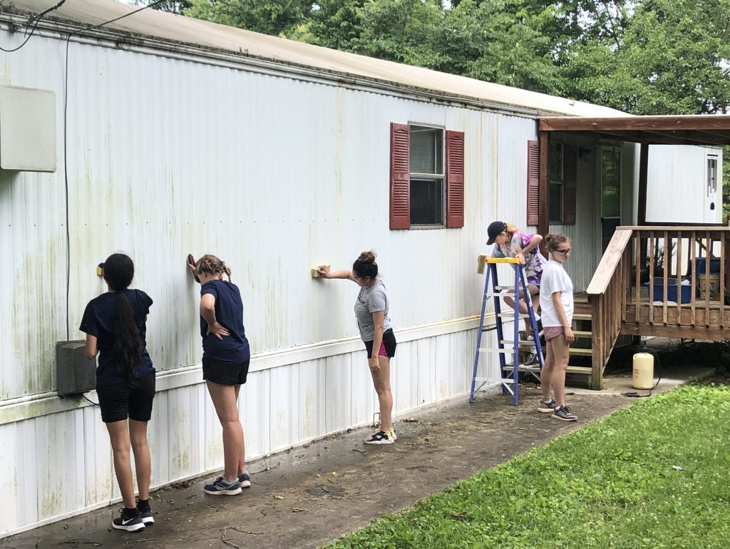 Five young women with scrub brushes work on the side of a white trailer home.