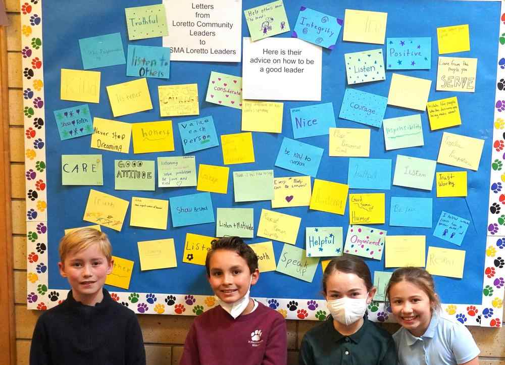 Four young kids smiling in front of a colorful poster of letters in uniforms.