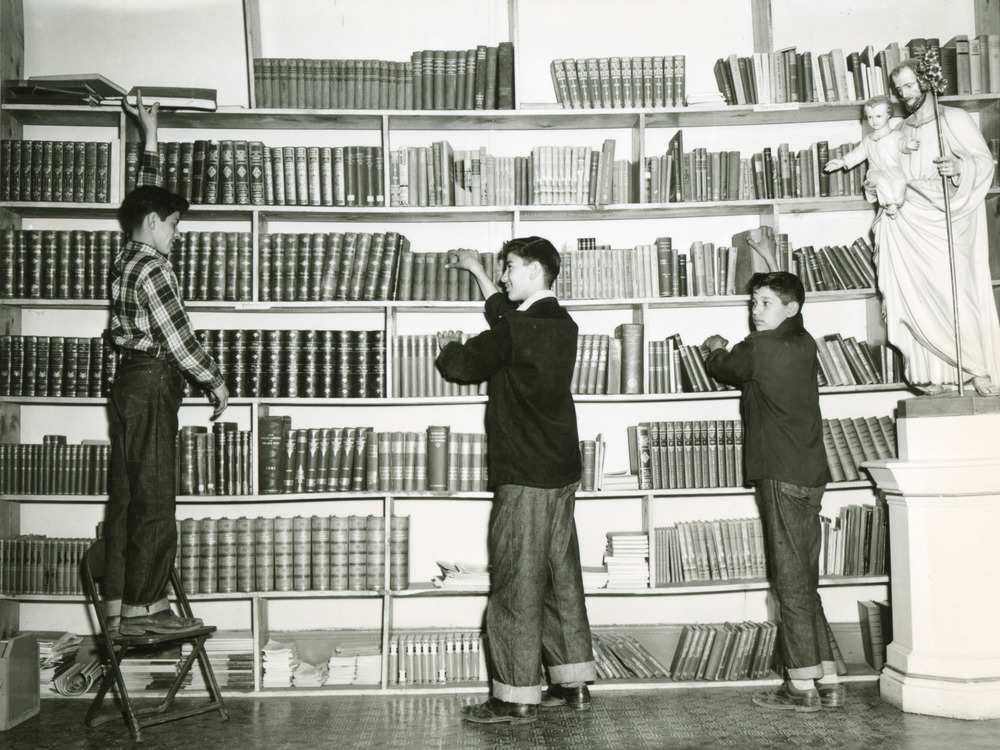 Archival photo of three boys standing in front of a wall of books. A statue of St. Joseph holding the infant Jesus is on the right.