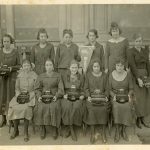Archival class photo of ten girls in early 20th century dresses. The five girls seated in the front row hold typwriters on their laps.