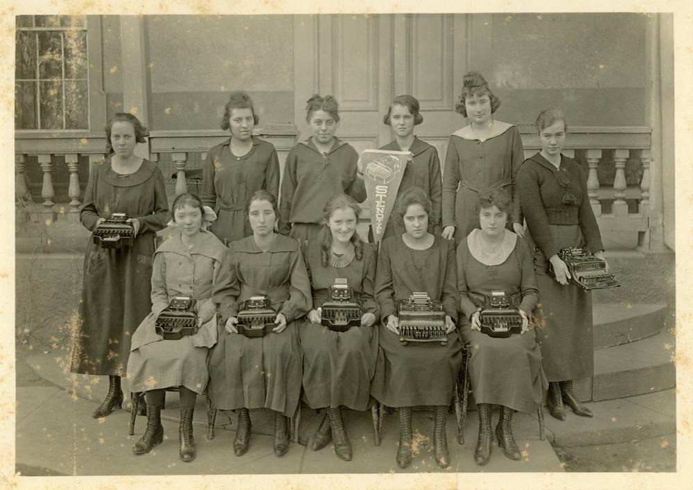 Archival class photo of ten girls in early 20th century dresses. The five girls seated in the front row and the two standing on the ends of the back row hold typewriters.