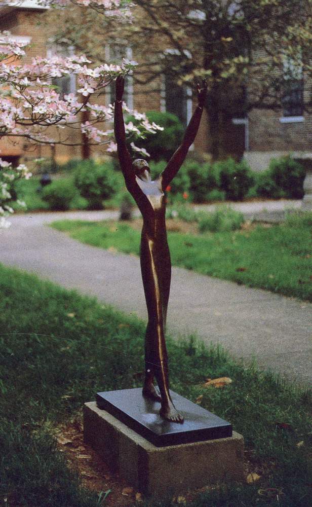 A bronze sculpture of a woman looking up and stepping forward with her hands reaching up to the sky. Trees flower behind her.