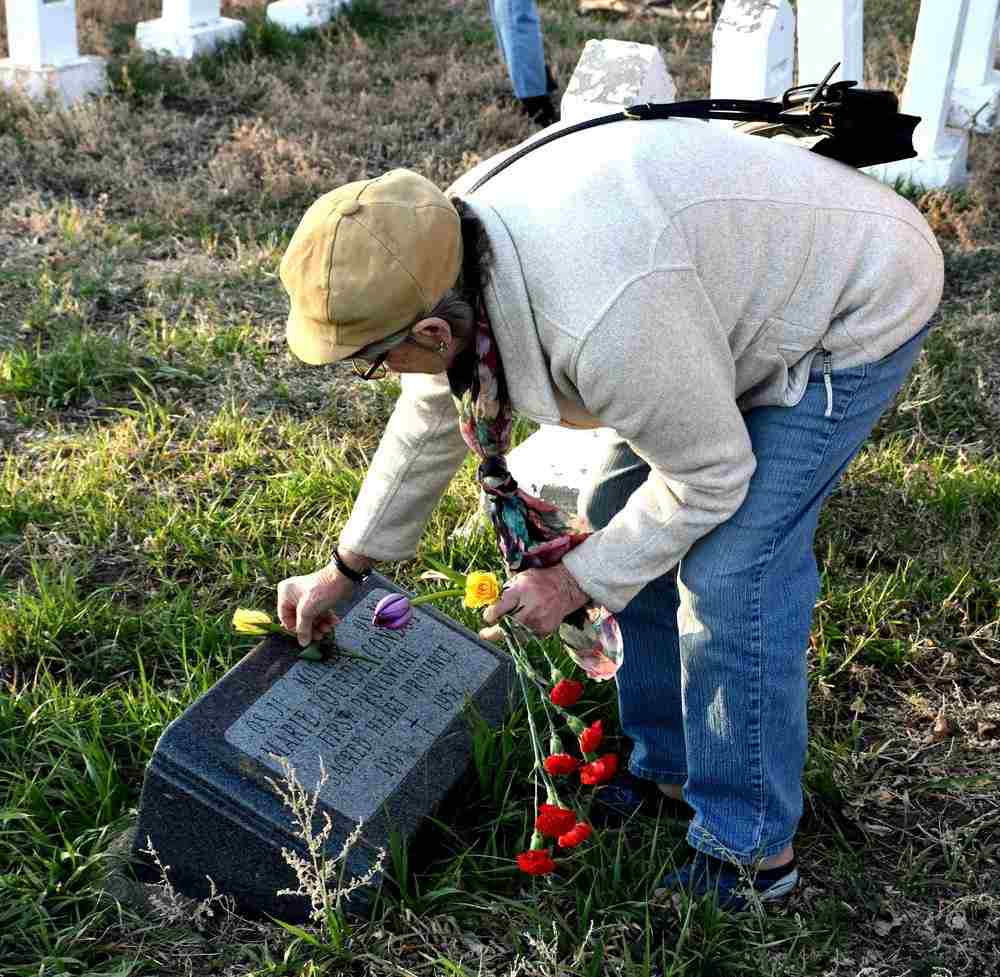 A woman wearing a brimmed cap bends down to lay a tulip on a low headstone. 