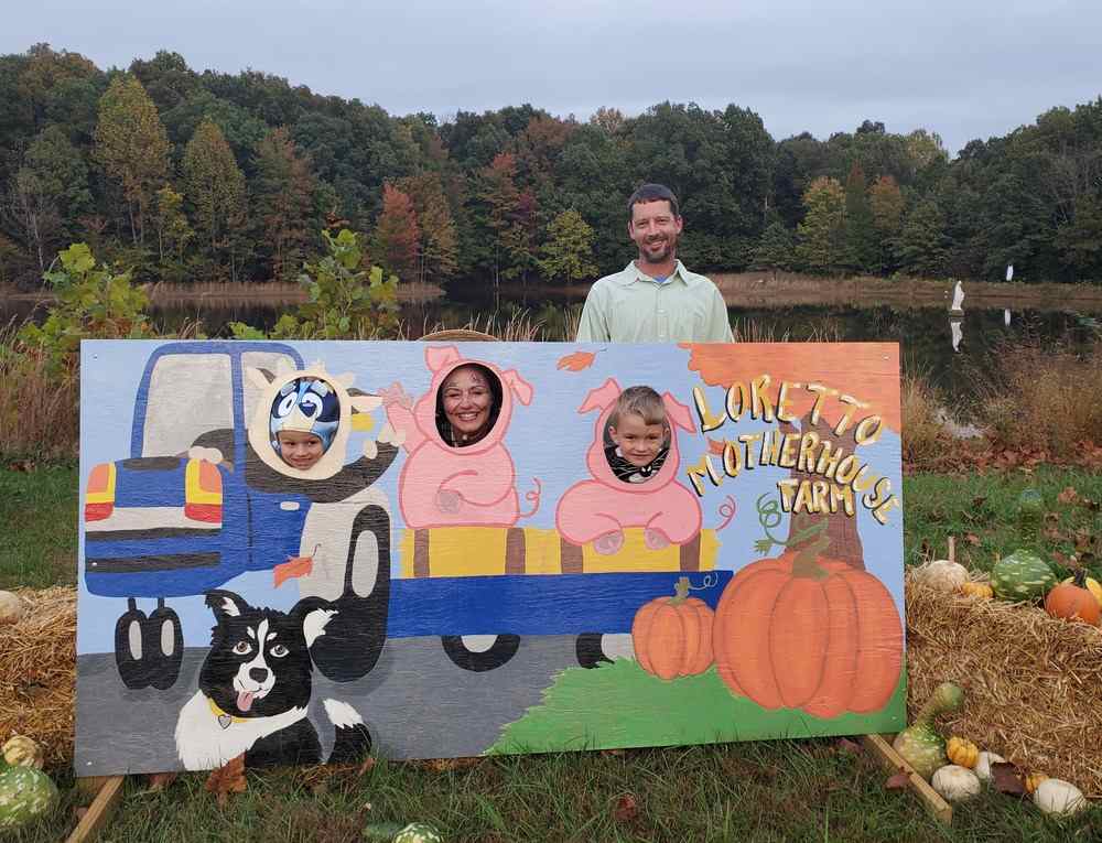 A woman and two children smile through holes in a photo op cutout drawing of a cow driving a tractor, pulling two pigs riding on the hay bales. The right side of the board reads "Loretto Motherhouse Farm." A smiling man stands behind the board.