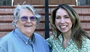 Two women posing and smiling brightly for a photo in front of a staircase.
