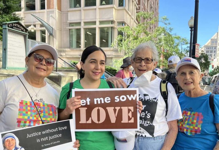 Four women stand together outside of St. Patrick's Church. Two hold signs reading "Justice without love is not justice" and "Let me sow love."