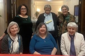 Six women sitting and standing together smiling for a photo while conversing.