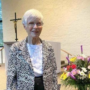 Bernie Feeney, a woman with short white hair, glasses and black and white dress clothes smiling for a picture in front of an indoor church stage next to a large vase of colorful flowers.