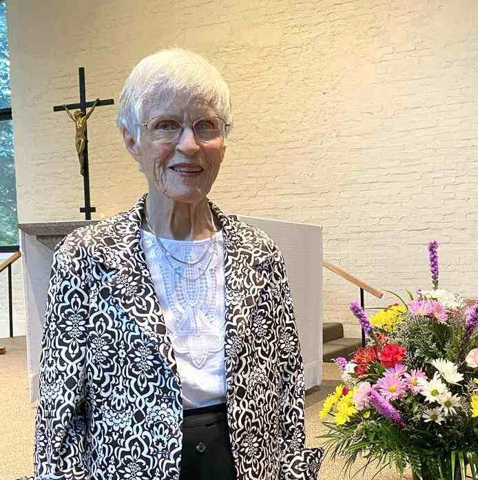 Bernie Feeney, a woman with short white hair, glasses and black and white dress clothes smiling for a picture in front of an indoor church stage next to a large vase of colorful flowers.