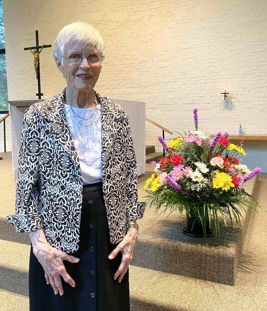 Bernie Feeney, a woman with short white hair, glasses and black and white dress clothes smiling for a picture in front of an indoor church stage next to a large vase of colorful flowers.