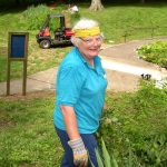 Woman with short, white curly hair and a yellow bandana pauses while weeding to smile for a photo.