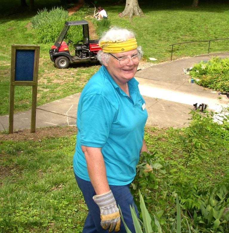 Woman with short, white curly hair and a yellow bandana pauses while weeding to smile for a photo.