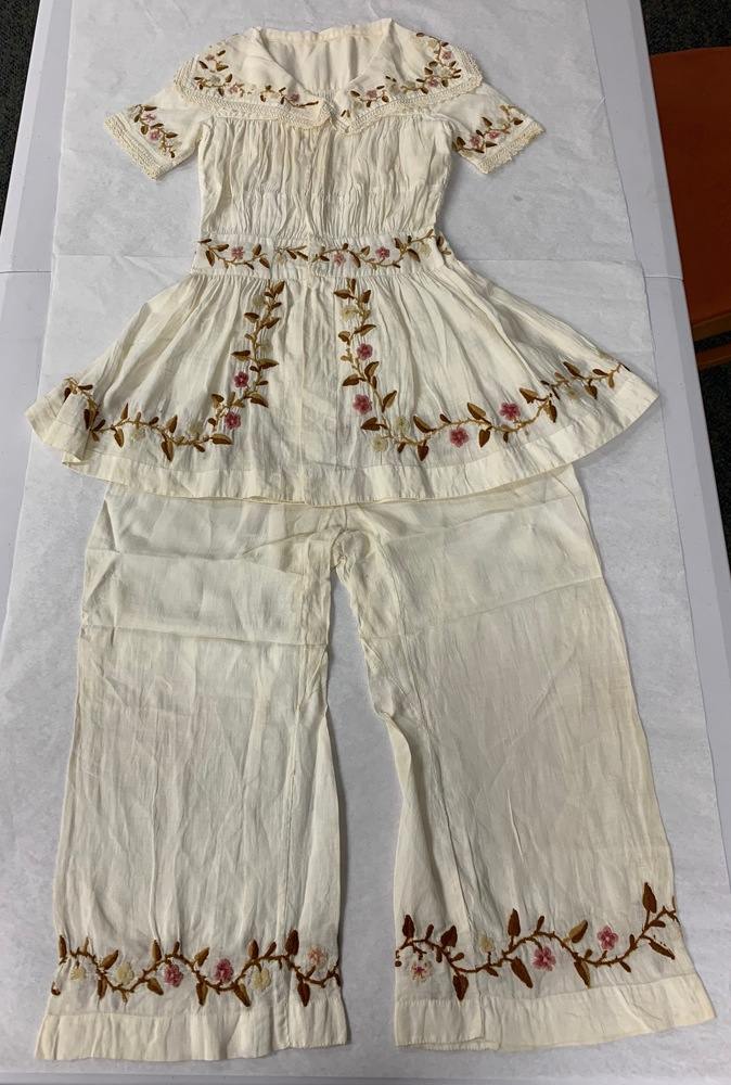 Photo of outfit for a young child - white pants with a large-collared tunic top. Both pieces are embroidered on the edges with vined leaves and flowers.