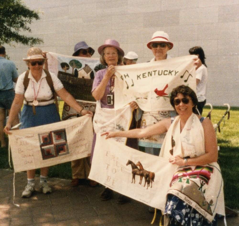 In this 1985 photo, five women wearing hats and/or sunglasses sit and stand together, holding up banners.