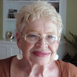 A woman with short white hair and oval-shaped glasses wearing a burnt orange shirt and white dangly earrings smiling brightly for a headshot picture indoors.
