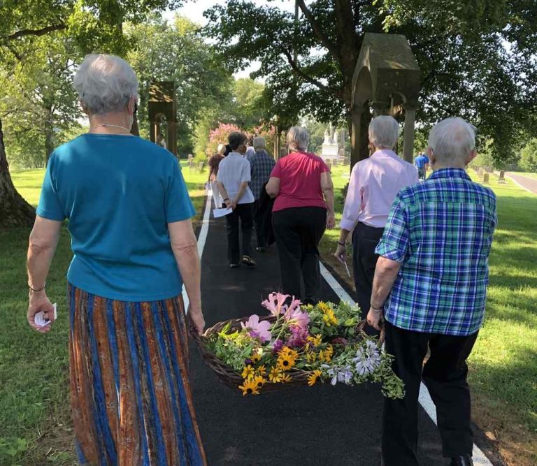 People process down a paved path on a green, summery day. The last two women in the processing carry a large basket of flowers between them.
