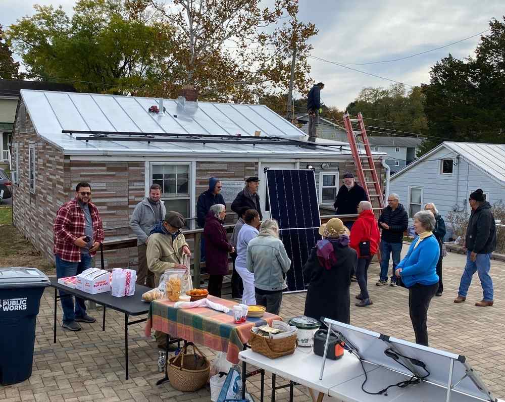 People in coats and warm hats gather behind a building with a ladder and solar panels. Card tables with food to share are in the foreground.