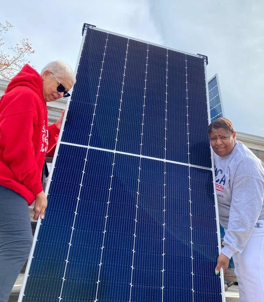 Two people team up to lift a very large solar panel up to the roof.