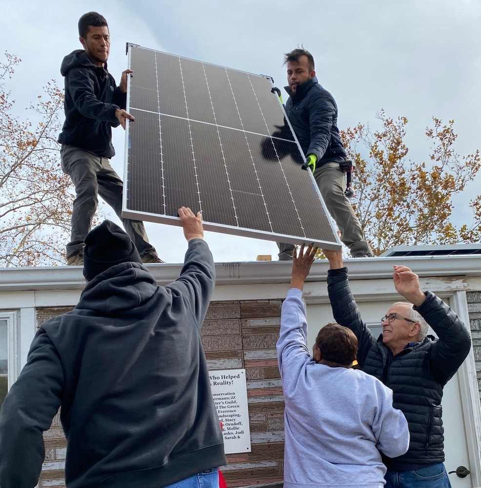 Five people, two on the roof of a one-story building and three on the ground, hoist a solar panel up to be installed on the roof.