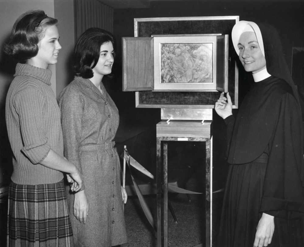 Archival photo of a nun in habit standing next to artwork on display. Two young women in 1960's fashion stand on the other side of the display.