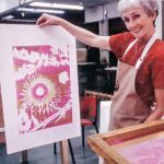 A smiling woman stands in a silkscreening workshop, holding up a beautiful print of pinks, reds and white.
