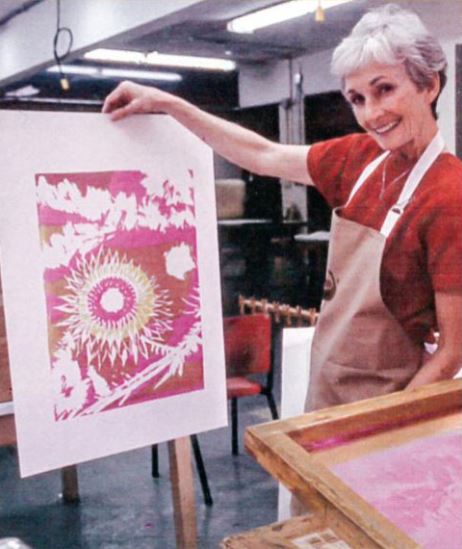 A smiling woman stands in a silkscreening workshop, holding up a beautiful print of pinks, reds and white.