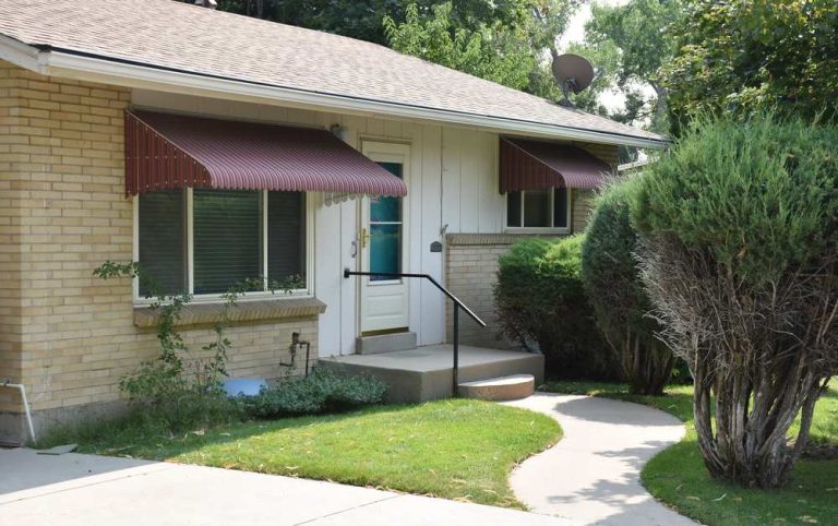 Osceola House, a beige brick, one-story with dark red window awnings and a brown shingle roof with a green grass lawn, concrete driveway, and large green bushes.