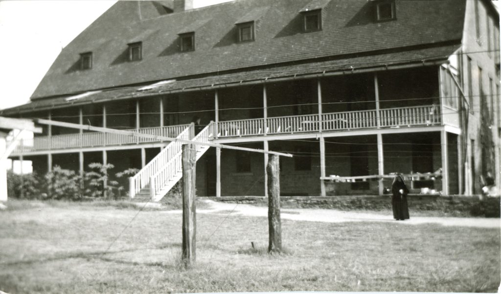Archival photo of large two story building with a balcony running the entire length of the second floor. A habited nun is standing in front of the building.