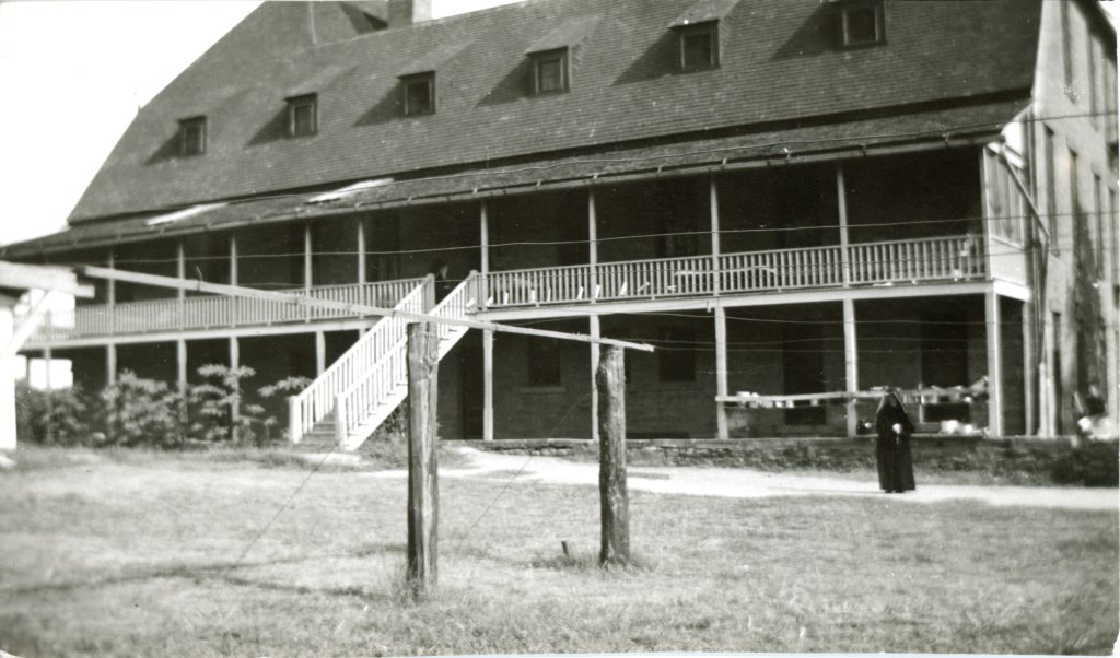 Archival photo of large two story building with a balcony running the entire length of the second floor. A habited nun is standing in front of the building, next to a clothesline.