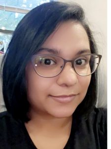 A woman with short black hair and dark-framed glasses smiling softly for a selfie indoors. She is showing off her new, short haircut after donating her hair.