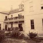 Indian School Archived Image: A black and white image of a large light-colored stone boarding school with a two-story balcony and deck filled with numerous students in dark-colored long dress uniforms on both the upper and lower levels of the balcony and deck.