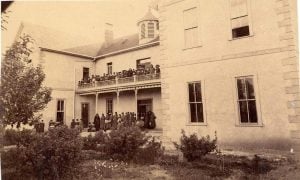 Indian School Archived Image: A black and white image of a large light-colored stone boarding school with a two-story balcony and deck filled with numerous students in dark-colored long dress uniforms on both the upper and lower levels of the balcony and deck.