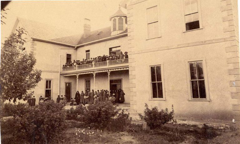 Indian School Archived Image: A black and white image of a large light-colored stone boarding school with a two-story balcony and deck filled with numerous students in dark-colored long dress uniforms on both the upper and lower levels of the balcony and deck.