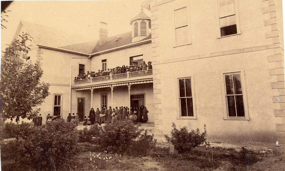 Indian School Archived Image: A black and white image of a large light-colored stone boarding school with a two-story balcony and deck filled with numerous students in dark-colored long dress uniforms on both the upper and lower levels of the balcony and deck.